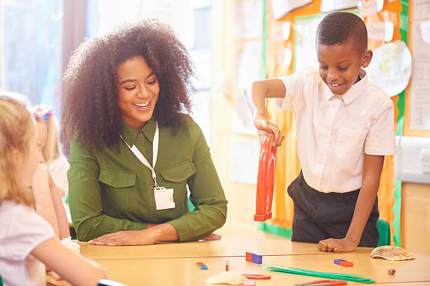 primary aged schoolchildren sit at their desks and play with various magnets as they discover science with their teacher .